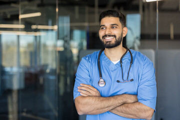 Fototapeta premium Confident young male doctor or nurse wearing blue scrubs and a stethoscope with arms crossed, smiling while looking away, representing dedicated healthcare professionals