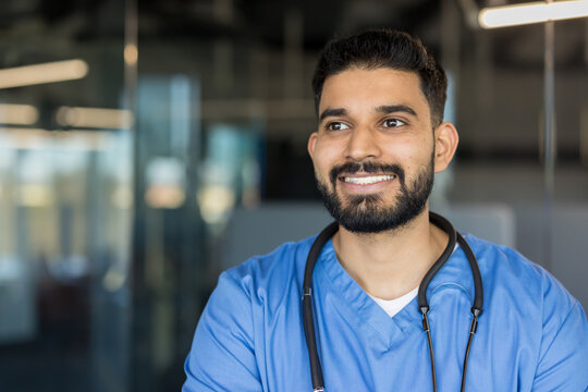 Young male doctor or medical professional with a beard smiling, looking away with a thoughtful or hopeful expression, wearing blue scrubs and a stethoscope in a modern clinic or hospital setting