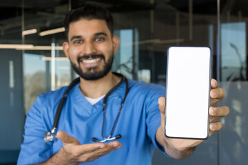 Smiling male doctor wearing blue scrubs and a stethoscope, holding up a smartphone with a blank white screen, offering a mockup space for healthcare applications
