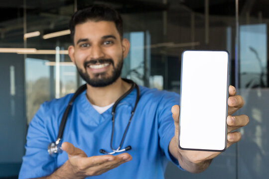 Smiling male doctor wearing blue scrubs and a stethoscope, holding up a smartphone with a blank white screen, offering a mockup space for healthcare applications - Powered by Adobe