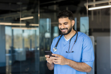 Indian male doctor wearing blue scrubs and a stethoscope, smiling while checking or messaging on his cellphone, representing modern healthcare communication and technology