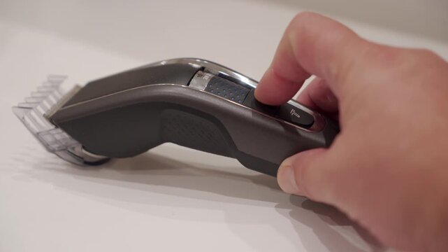 A hand pushes the plastic power switch on a cordless hair clipper. The electric appliance instantly begins working, emitting a loud, buzzing, and whirring sound.