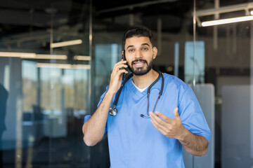 Young indian male doctor in blue scrubs and stethoscope having an animated conversation on a mobile phone, discussing patient care in a modern hospital setting