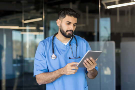 Indian male doctor in blue scrubs with stethoscope, focused on reviewing patient data on a tablet in a bright modern clinic, symbolizing digital healthcare and telemedicine