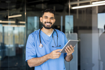 Bearded male nurse wearing blue scrubs and stethoscope, standing confidently while holding a digital tablet, representing modern healthcare technology and patient care