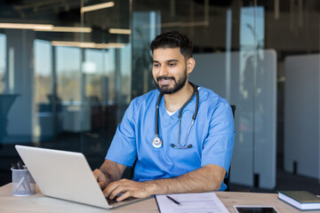 Indian male doctor wearing blue scrubs and stethoscope working on a laptop at his office desk,...