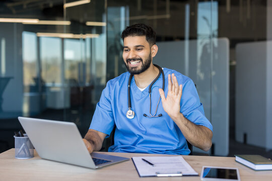 Male professional doctor wearing blue scrubs and stethoscope smiling, waving hand during virtual teleconsultation from an office, providing remote healthcare services to patients