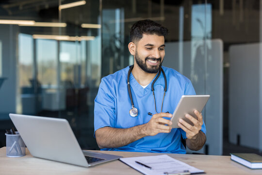 Indian male doctor or nurse wearing scrub uniform and stethoscope smiling while reviewing medical ,information on a digital tablet at a modern clinic desk with a laptop