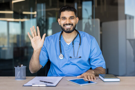 Male healthcare professional wearing scrubs and a stethoscope, smiling and waving at the camera from his desk in an office setting, offering friendly medical support remotely