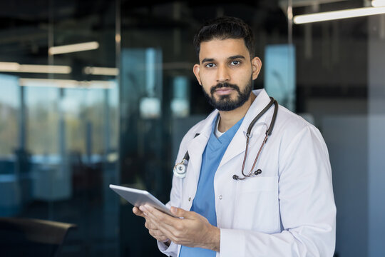Young indian man working as a doctor wearing a white lab coat and stethoscope, holding a digital tablet while standing in a modern clinic or hospital setting