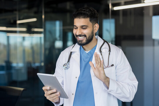 Young male doctor in a lab coat and stethoscope is smiling, waving during a telehealth video call on a digital tablet, offering remote medical advice and online consultation warmly