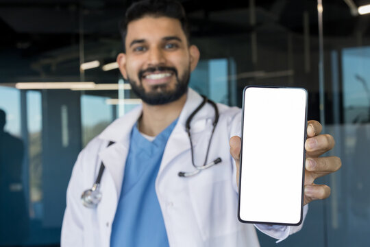 Smiling professional indian doctor wearing a lab coat and stethoscope showing a modern smartphone with a blank white screen, symbolizing digital healthcare and online consultations - Powered by Adobe