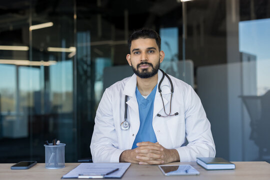 Confident indian male doctor in white coat with stethoscope, sitting at a modern clinic desk, looking calmly at camera, conveying professionalism, expertise and patient care