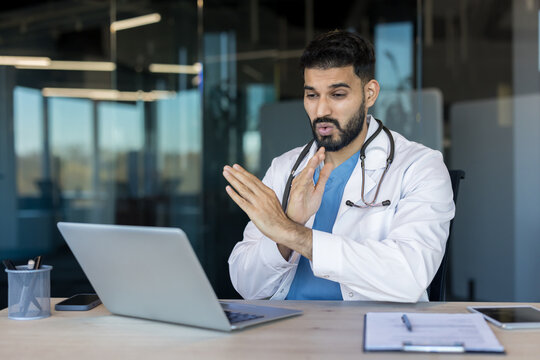 Indian doctor wearing a lab coat and stethoscope consulting a patient virtually using a laptop, providing remote healthcare services from a modern office setting - Powered by Adobe