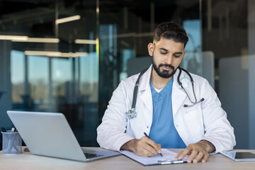 Young male physician wearing a lab coat and stethoscope working on healthcare documentation at a...