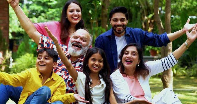 Indian family sitting on lawns smiling at camera, three generations including grandparents parents and children bonding together outdoors in park, happy Asian family enjoying leisure time together