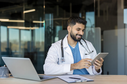 Young male doctor in white coat and stethoscope sits at desk, smiling while reviewing patient data on a digital tablet, representing modern telehealth and medical technology care