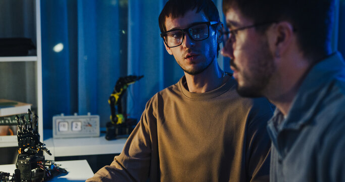Team adult male engineer collaborate on robotic hand project using AI code solutions machine learning at tech workspace night. Artificial intelligence interface, collaboration prototype development.