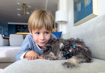 Joyful little boy hugging his schnauzer puppy dog lying on the couch at home.