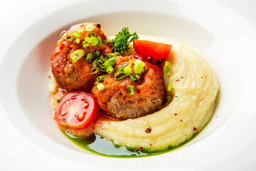 Two appetizing meatballs in tomato sauce served on creamy mashed potatoes, garnished with green onions and cherry tomatoes in a white bowl