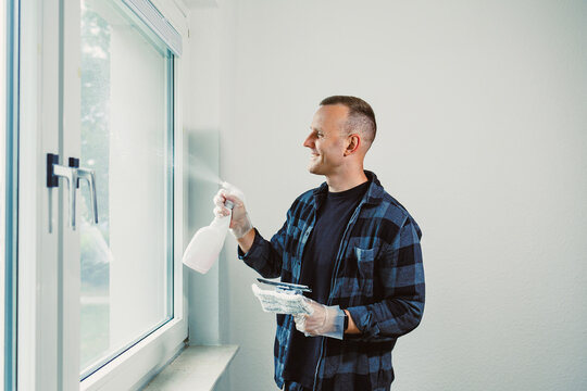 Man cleaning a window with a spray bottle and cloth in a sunlit room, showcasing a moment of satisfaction and cleanliness
