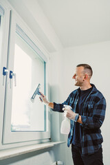 Man cleaning window with a squeegee in a bright room, enjoying the task while wearing gloves and a plaid shirt