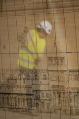 Safety screen texture filters the light showing a building worker wearing a yellow reflective vest working near detailed technical architectural drawings on the job sit