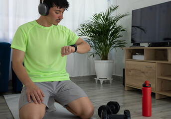 Man checking his smartwatch while training and listening to music with headphones in his living room. Concept of fitness technology, time tracking, and well being