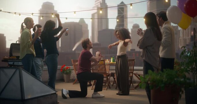Romantic Rooftop Proposal Unfolds as a Man Kneels, Offering a Ring to a Woman, Love of His Life. Friends Capture the Joyous Moment, Celebrating Their Engagement Under City Lights.