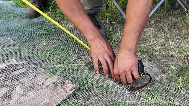 A man's hands using a yellow tape measure to check the dimensions of a metal mesh.