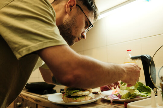 Focused man preparing homemade burger with fresh vegetables and lettuce in cozy kitchen. Concept of cooking, lifestyle, and food preparation