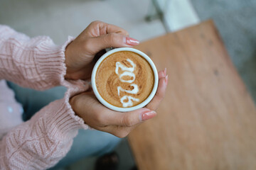 A person holding a cup of coffee with latte art showing the number "2026," wearing a pink sweater and relaxing indoors.