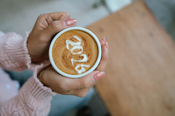 A person holding a cup of coffee with latte art showing the number "2026," wearing a pink sweater and relaxing indoors.