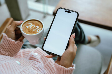 A person in a cozy pink sweater holding a cup of coffee with "2026" latte art while using a smartphone with a blank screen, symbolizing a fresh start for the new year.