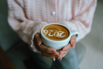 A person holding a cup of coffee with latte art showing the number "2026," wearing a pink sweater and relaxing indoors.