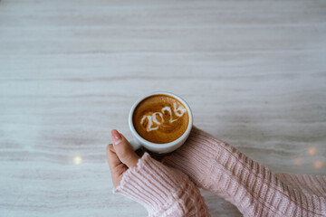A person holding a cup of coffee with latte art showing the number "2026," wearing a pink sweater and relaxing indoors.