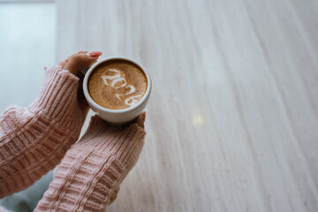 A person holding a cup of coffee with latte art showing the number "2026," wearing a pink sweater and relaxing indoors.