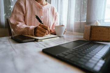 Woman using smartphone at cozy workspace with laptop, notebook, and coffee cup on marble table in soft daylight atmosphere.