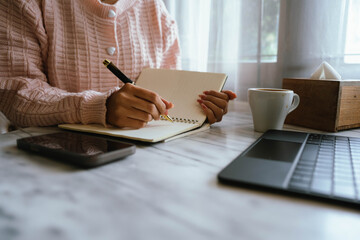 Woman using smartphone at cozy workspace with laptop, notebook, and coffee cup on marble table in soft daylight atmosphere.