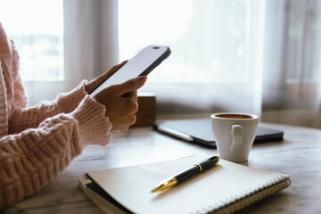 Woman using smartphone at cozy workspace with laptop, notebook, and coffee cup on marble table in soft daylight atmosphere.