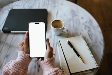 Woman using smartphone at cozy workspace with laptop, notebook, and coffee cup on marble table in soft daylight atmosphere.