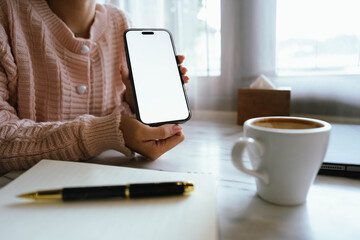 Woman using smartphone at cozy workspace with laptop, notebook, and coffee cup on marble table in soft daylight atmosphere.