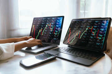 A person holding a smartphone showing stock market data with trading charts displayed on laptop screens in the background.