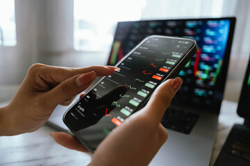 A person holding a smartphone showing stock market data with trading charts displayed on laptop screens in the background.