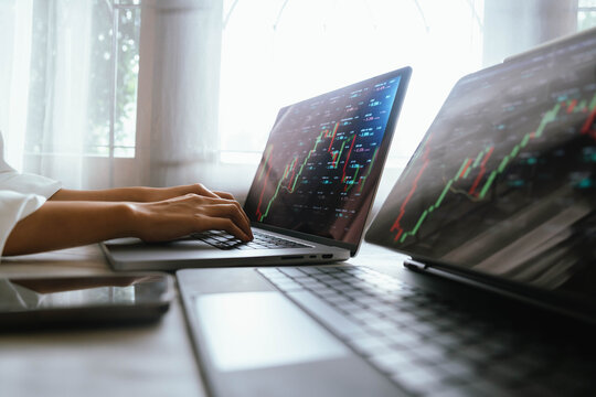 A person holding a smartphone showing stock market data with trading charts displayed on laptop screens in the background. - Powered by Adobe