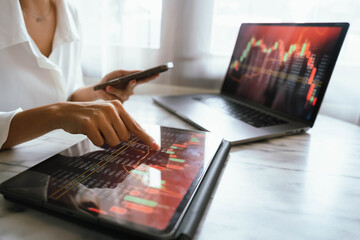 A person holding a smartphone showing stock market data with trading charts displayed on laptop screens in the background.