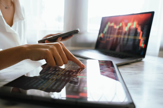 A person holding a smartphone showing stock market data with trading charts displayed on laptop screens in the background.