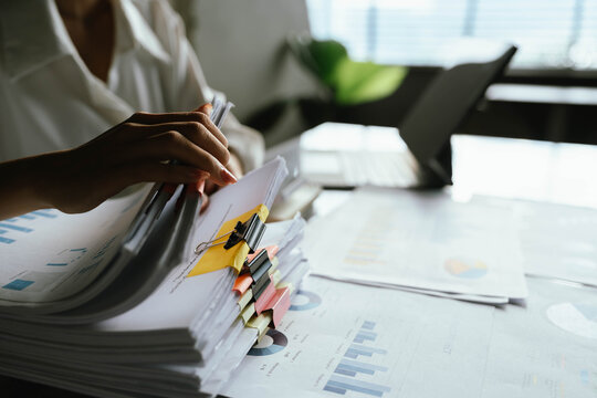 Close-up of businesswoman working at her desk with laptop and stack of documents, focusing on paperwork, reports and financial analysis in a busy office environment. - Powered by Adobe
