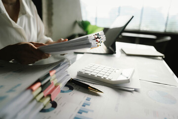 A person working with a calculator and financial documents on a desk, analyzing data from charts displayed on a laptop in a bright office environment.