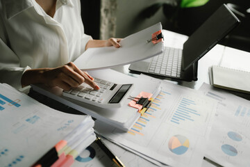 A person working with a calculator and financial documents on a desk, analyzing data from charts displayed on a laptop in a bright office environment.